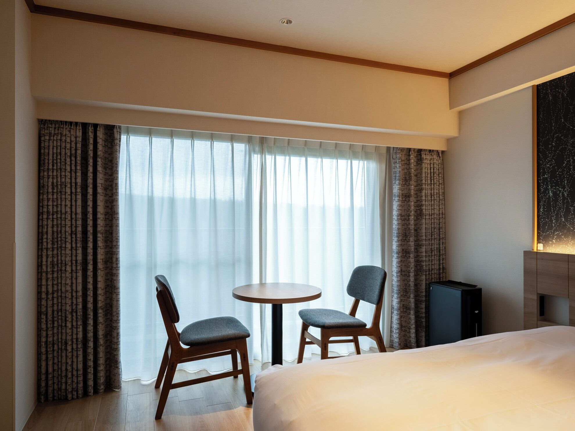 A hotel room with a bed, two chairs, a small table, and a large window with curtains.