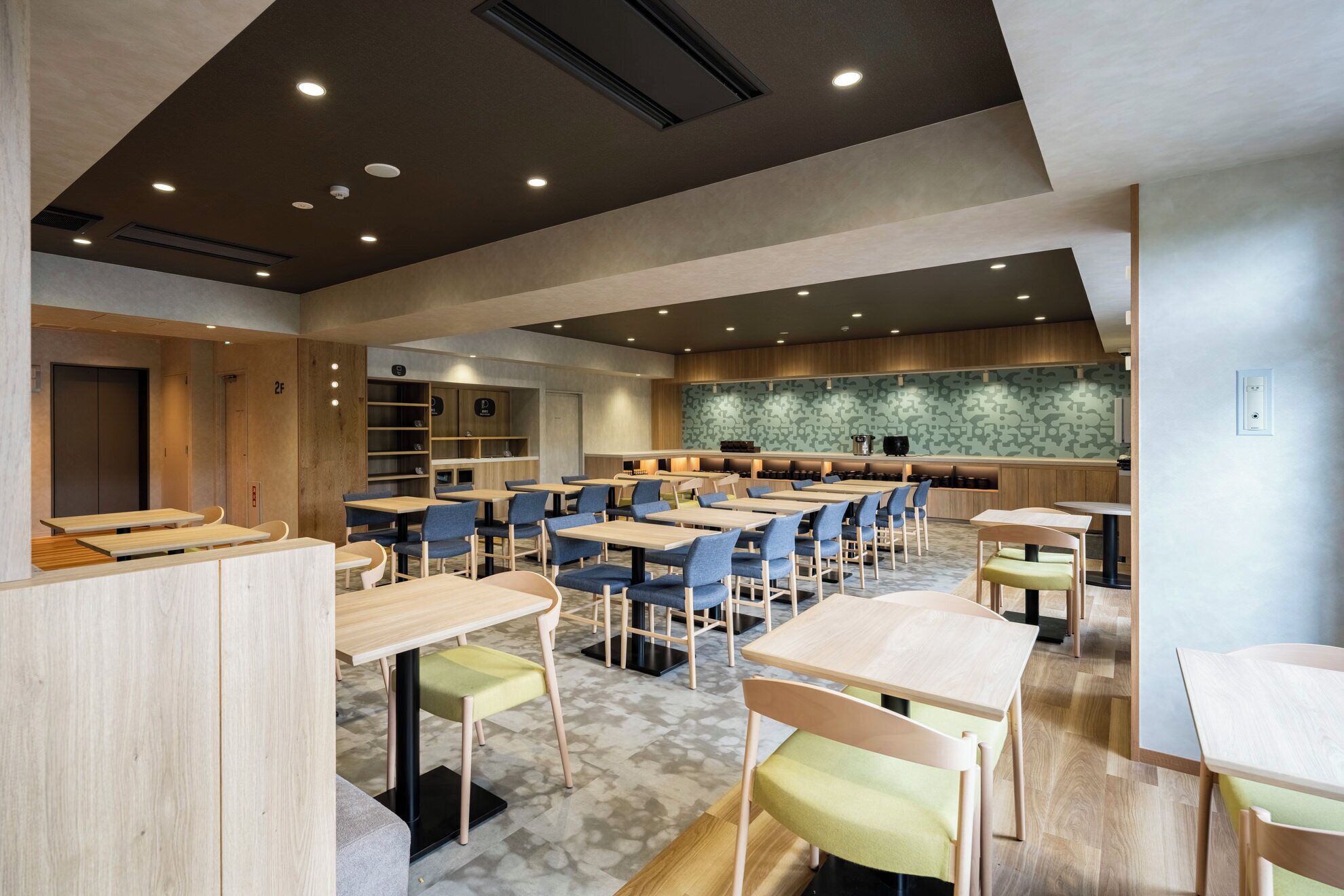 A modern dining area with multiple tables and chairs, a kitchen in the background, and a patterned wall.