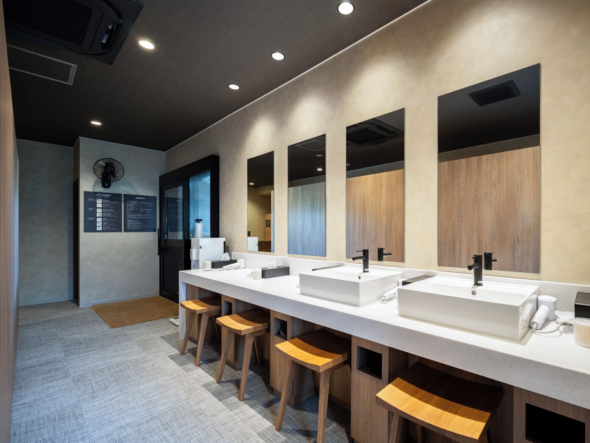 A modern bathroom with three sinks, wooden stools, and a fan.