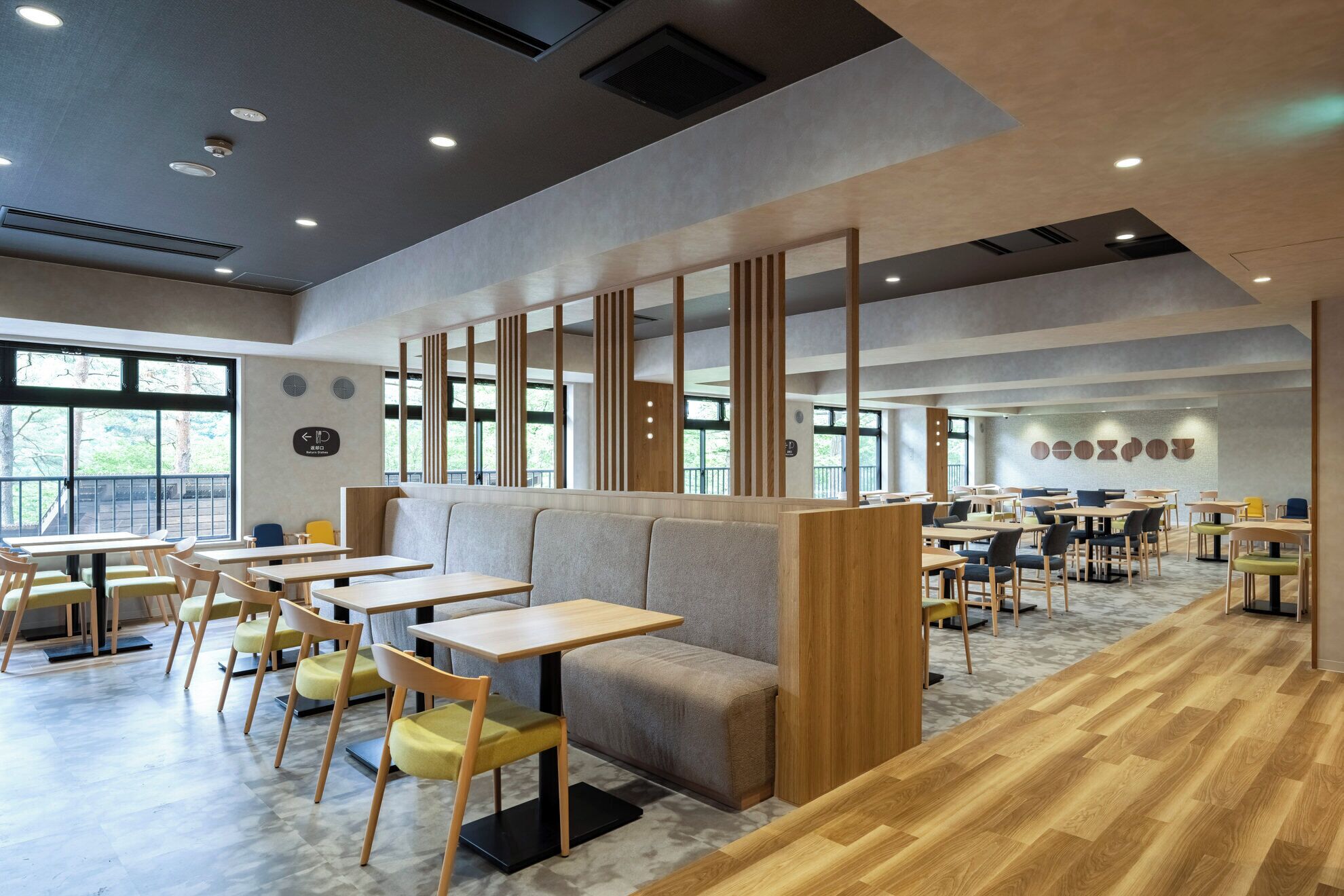 A modern restaurant with wooden tables and chairs, a central seating area, and large windows.