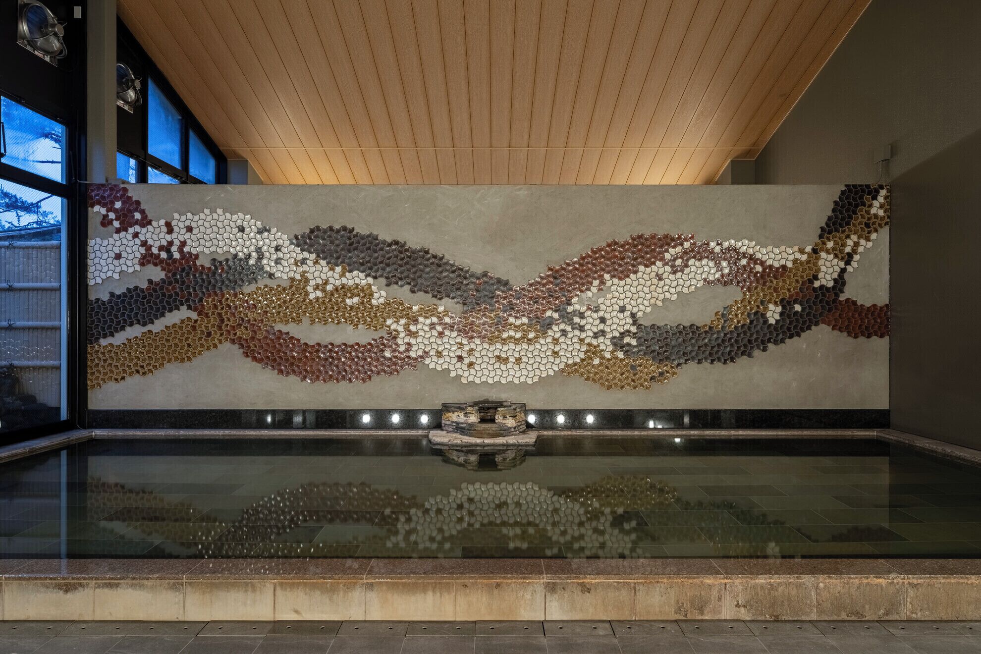A modern interior with a reflective water feature, wooden ceiling, and a wall with a mosaic pattern.