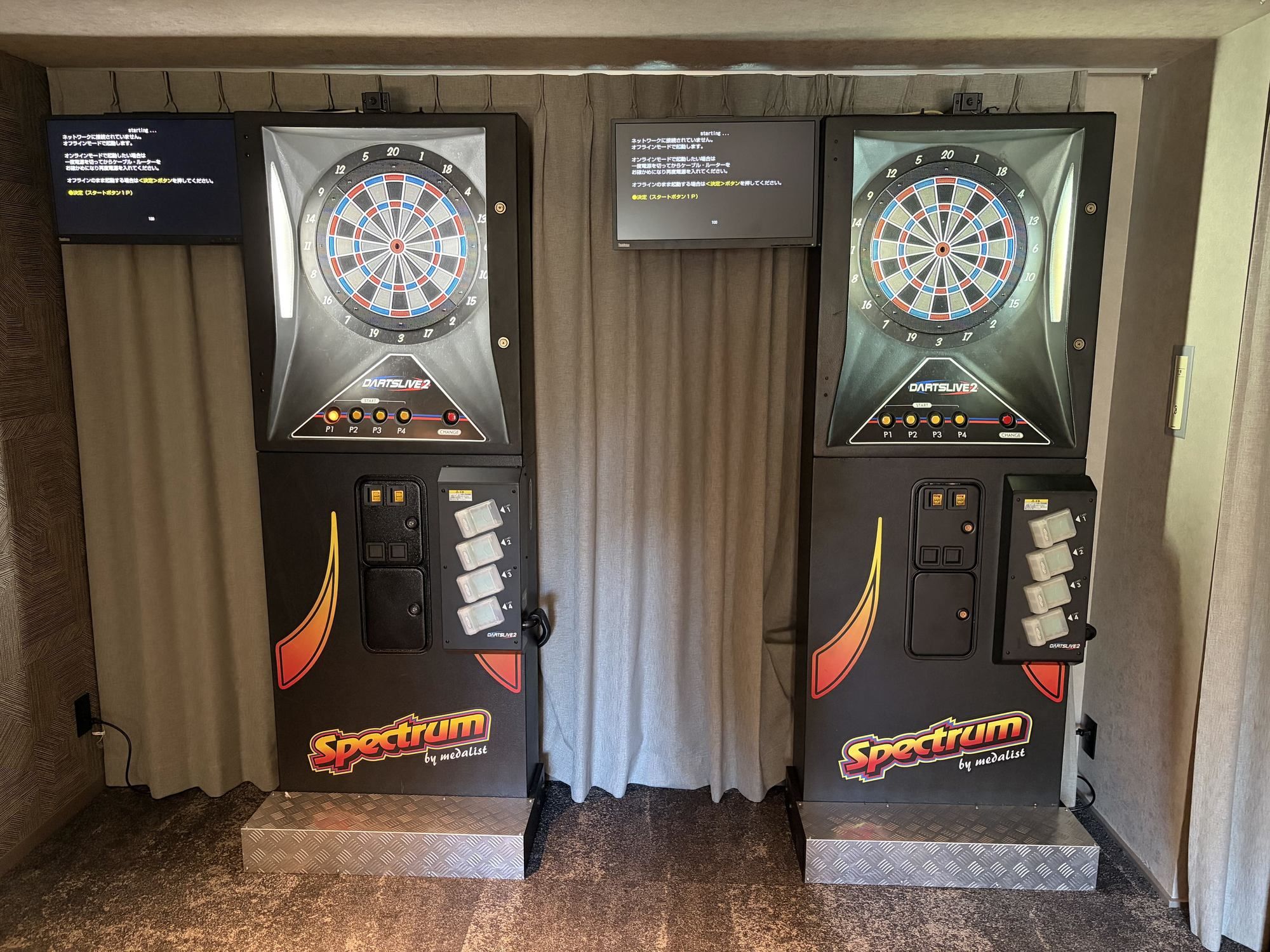 Kaksi dart-taulua, joissa on merkintä ”DartsLive2”, sekä pistetaulut ja ohjauspaneelit, ja taustalla verho.