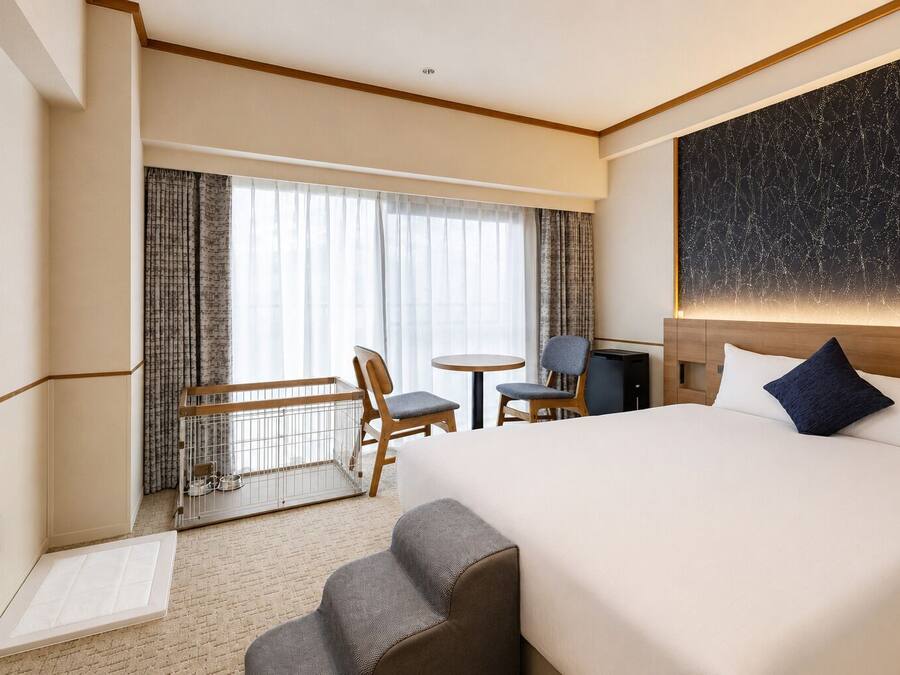 A hotel room with a large bed, a desk, two chairs, a small table, a balcony with a railing, and a large window with curtains.