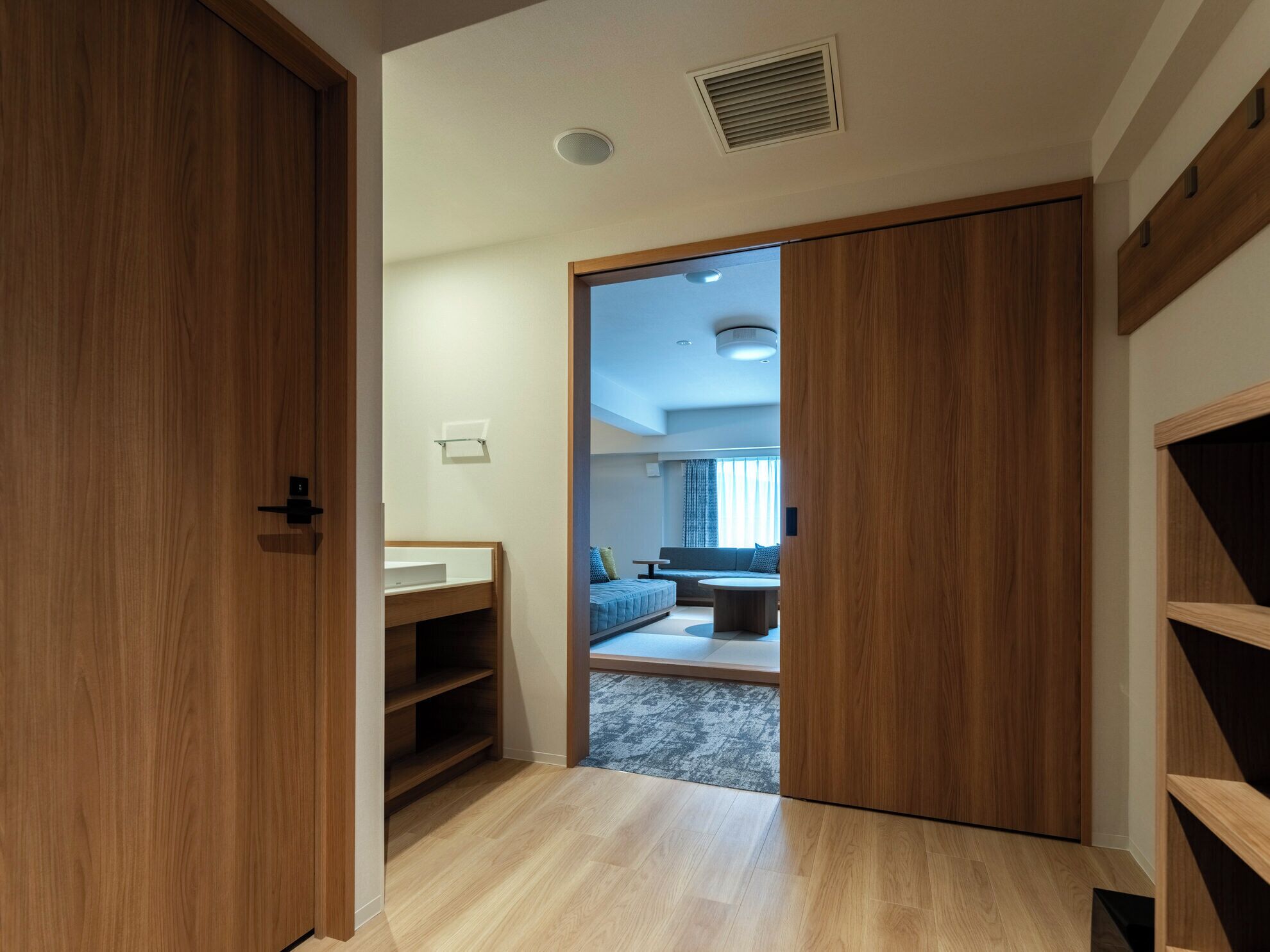 A modern hotel room with a wooden wardrobe, a built-in shelf, and a view into a living area with a sofa and a round table.