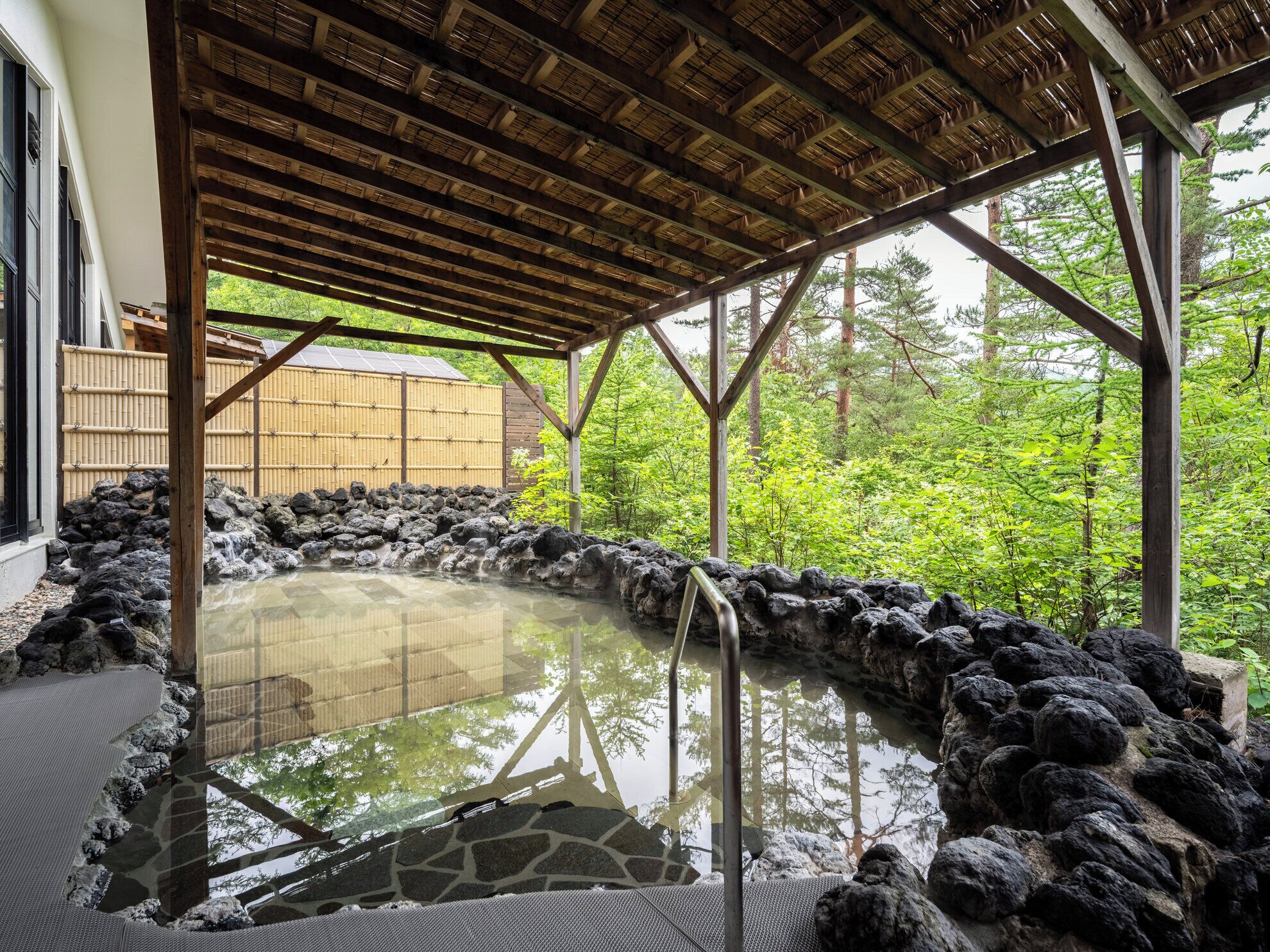 A hot spring area with a stone wall surrounding the water, wooden beams overhead, and a view of a forested area.