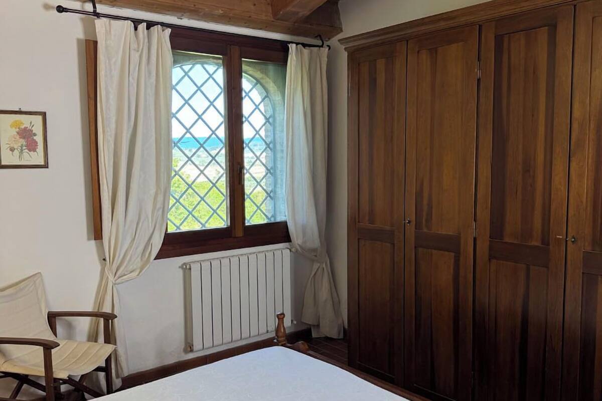 A bedroom with a wooden ceiling, a large bed, a window with curtains, a radiator, and a wooden wardrobe.