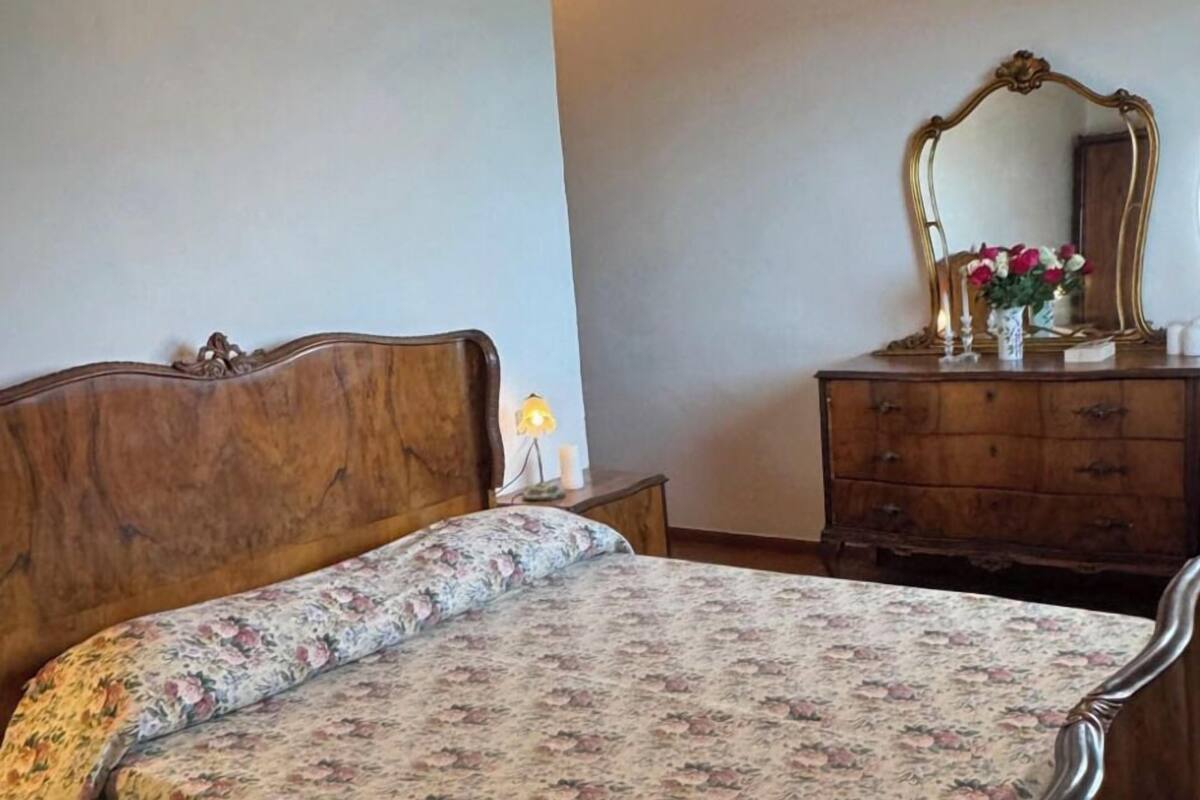 A bedroom with a wooden bed, a floral bedspread, a nightstand, a dresser, a mirror, and a wall-mounted light fixture.