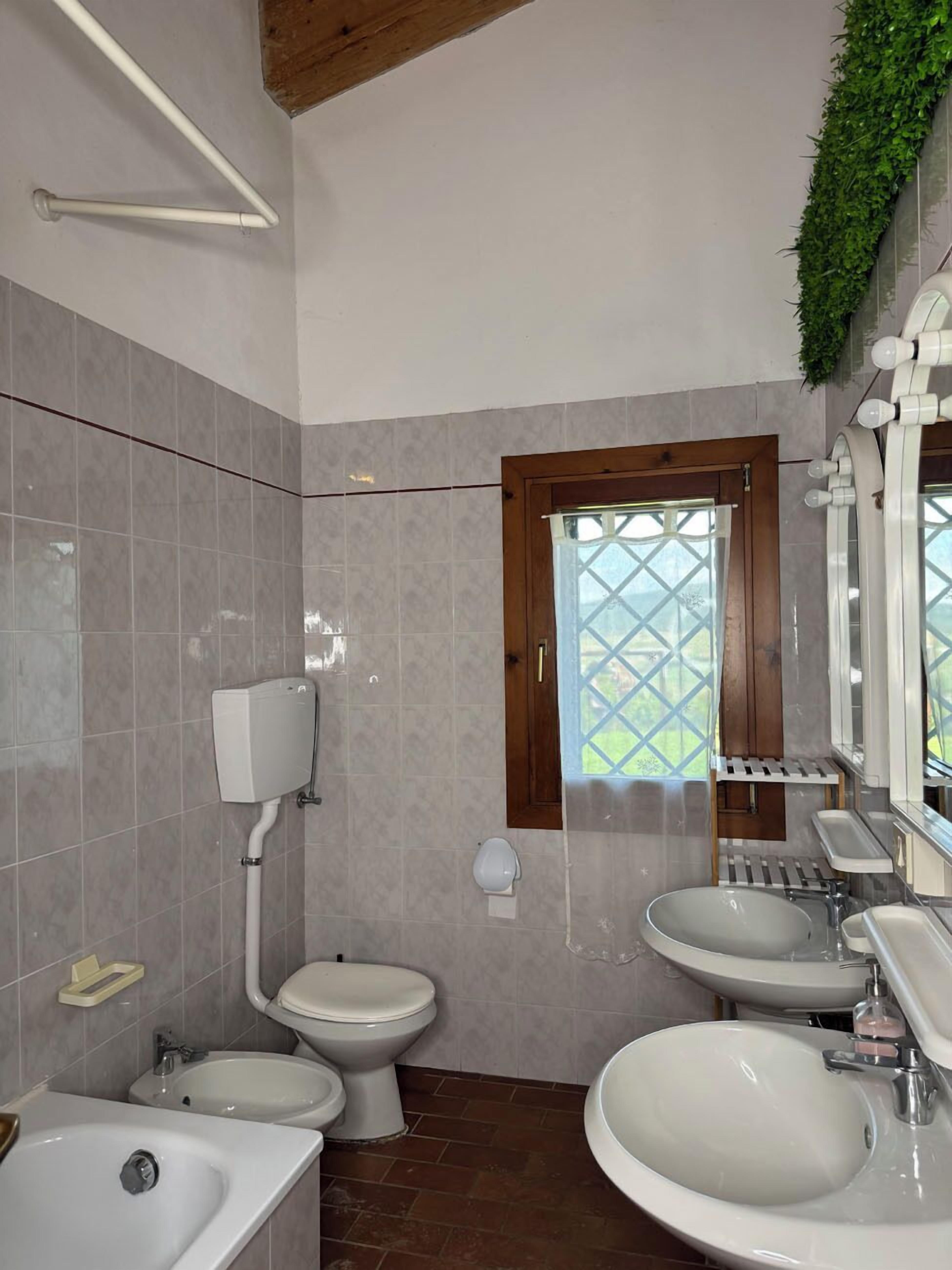Un baño con dos inodoros, dos lavamanos, un dispensador de papel higiénico, un ventanal con vistas y una zona de ducha.