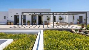 A modern building with a covered patio area, potted plants, and a landscaped garden with yellow flowers. - Villa Lemoni – Walk to the Beach (4798+R23 Xifara, Greece)