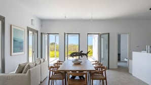 A modern dining area with a long table, wicker chairs, and a view of the outdoors. - Villa Lemoni – Walk to the Beach (4798+R23 Xifara, Greece)