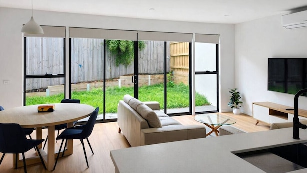 A modern living room with a dining area, large sliding glass doors to a backyard, a flat-screen TV mounted on the wall, and a minimalist design.