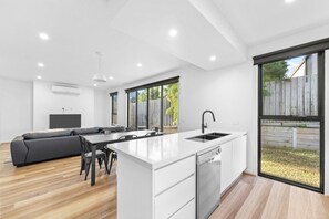 A modern kitchen with a central island, a dining area with a sofa, and a large window providing a view of the outdoors. - Sleek Contemporary Haven Geelong (Highton)
