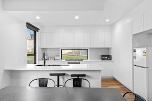 A modern kitchen with white cabinetry, a large island with seating, a built-in oven, and a view of the outdoors. - Sleek Contemporary Haven Geelong (Highton)