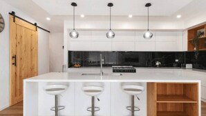 A modern kitchen with a large island, three bar stools, pendant lighting, and a wooden door. - Modern Luxe Haven – Your Geelong Escape (Highton)