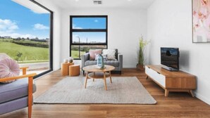 A modern living room with a sofa, coffee table, TV stand, and a large window offering a view of a green landscape. - Modern Luxe Haven – Your Geelong Escape (Highton)