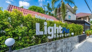 Un letrero para 'Light Plus Khon Lab' con techo de tejas rojas, exuberante follaje y una pared de concreto.