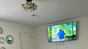 Una stanza con un letto, una TV a muro, una ventola a soffitto e una porta.