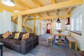 A spacious living area with a leather sofa, wooden beams, a staircase, a dining table, and a kitchen area. - Nature experience & close to the city of Berlin: "THE WELSH HOUSE" (No. 56a) (Schönwalde-Glien)