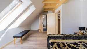 A bedroom with a sloped ceiling, skylight, wooden flooring, a bench, a bed with a patterned bedspread, and a nightstand with a lamp. - Nature experience & close to the city of Berlin: "THE WELSH HOUSE" (No. 56a) (Schönwalde-Glien)