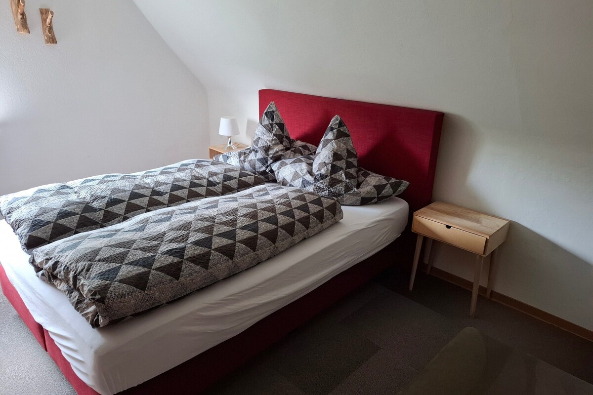 Een bed met een rode hoofdbord, beddengoed met een grijs en zwart patroon, en een klein houten nachtkastje met een lamp.