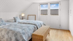 A bedroom with a sloped ceiling, two beds with patterned bedding, a bedside lamp, a wicker bench, and a view through a door. - 4 Star Holiday Home in Lokken (Lokken)
