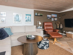 A modern living room with a sofa, a brown leather chair, a round coffee table, and a flat-screen TV mounted on the wall. - 4 Star Holiday Home in Hejls (Hejls)