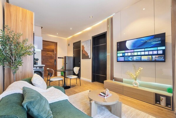 A modern hotel room with a sofa, a small table, a TV, a desk, and a plant. - Mildy Lodge And Apartment  (Abuja)