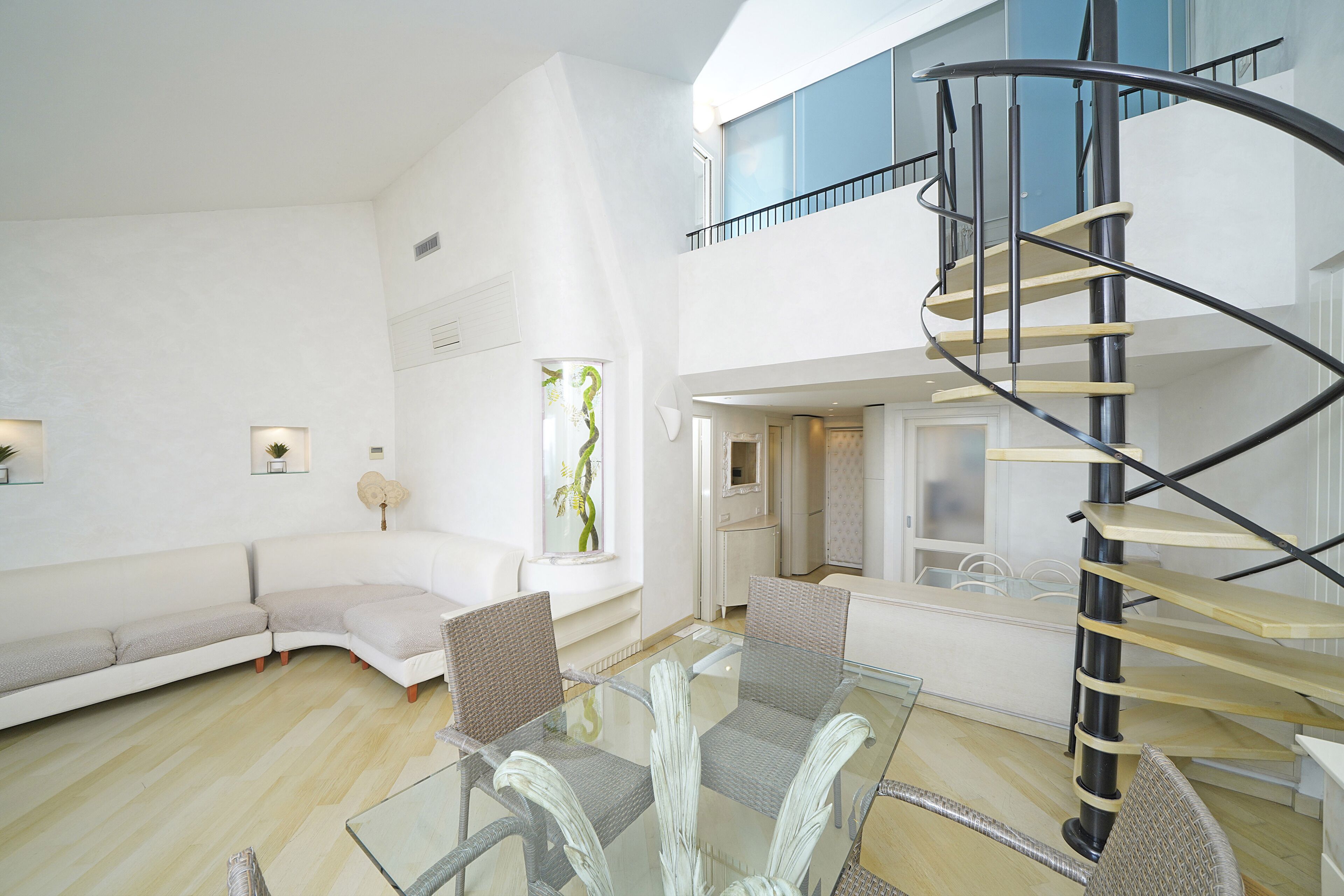 A modern living room with a glass table, wicker chairs, a white sofa, and a spiral staircase.