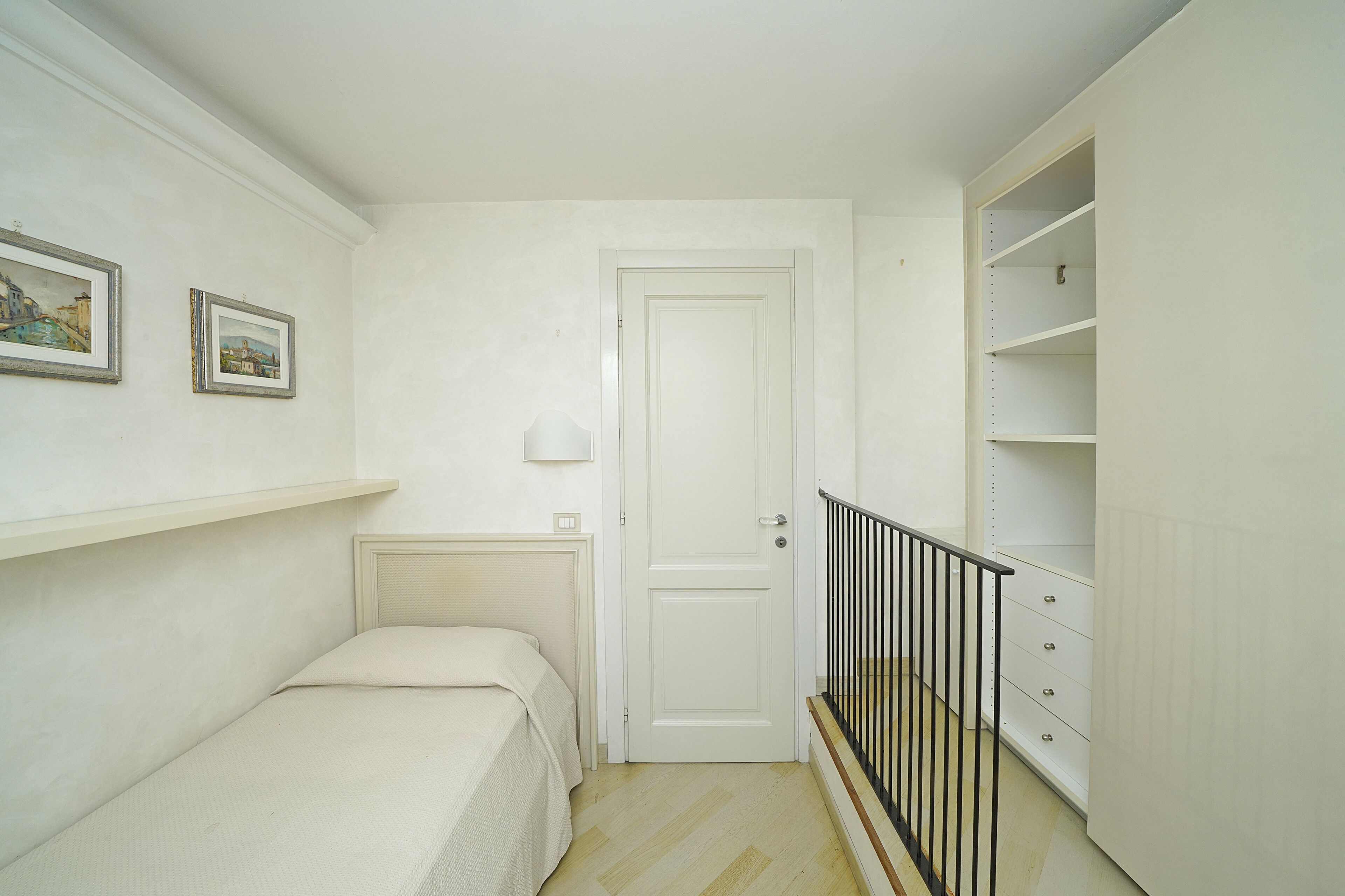 A bedroom with a bed, a door, a small shelf, and a closet with a black railing.
