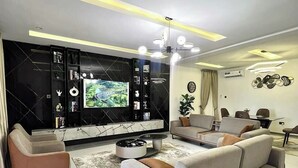 A modern living room with a large flat-screen TV, a sofa, a coffee table, and a dining area with a table and chairs. - House 21 Hotel (Asaba)