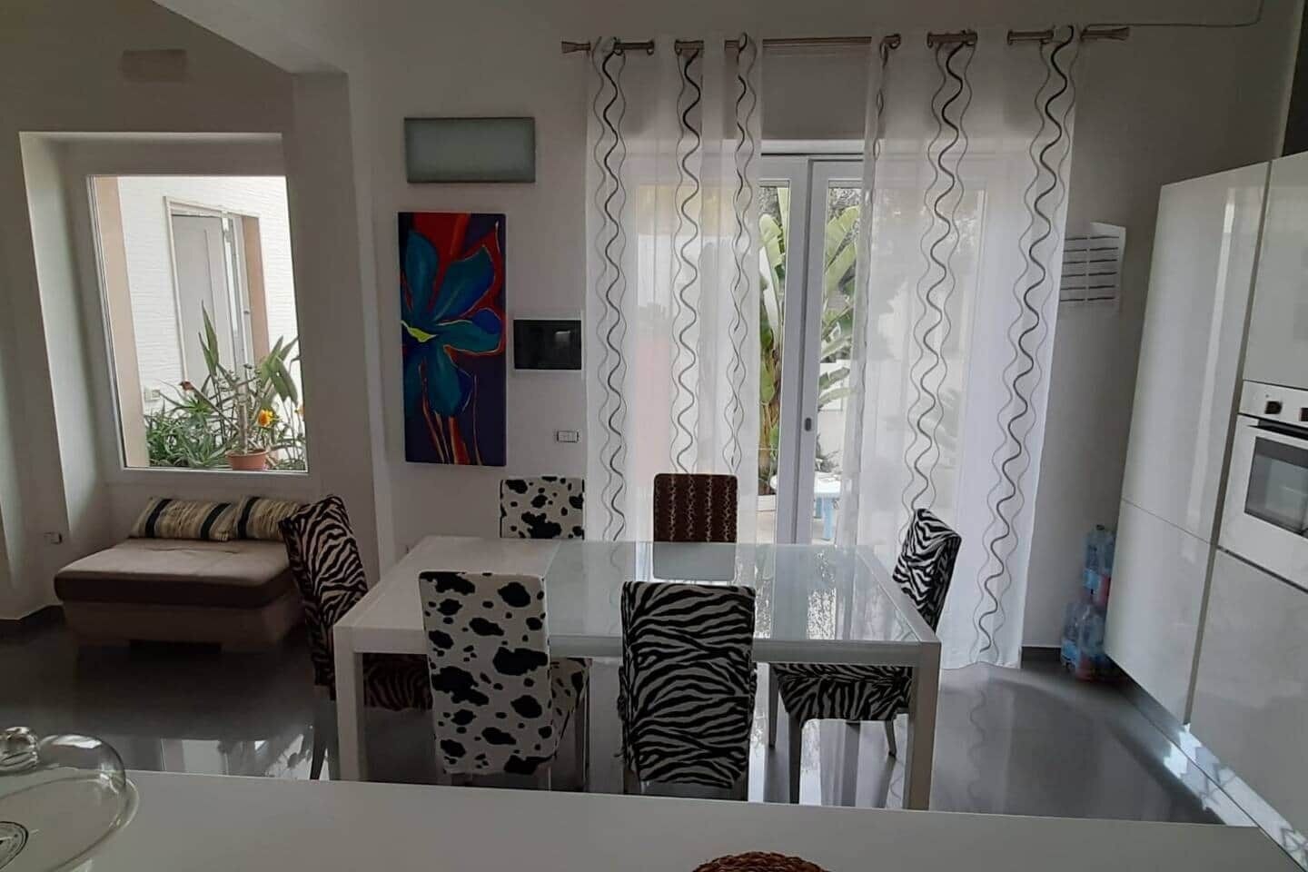 A modern kitchen with a refrigerator, a dining table with zebra-print chairs, a sofa with patterned cushions, and a large window with curtains.