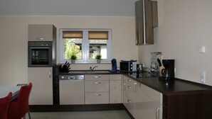 Eine moderne Küche mit Einbauofen, Mikrowelle und Kaffeemaschine. Ein Fenster mit Jalousien, ein Spülbecken und ein Essbereich mit einem Stuhl sind vorhanden.