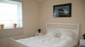 Une chambre avec un lit blanc, une table de chevet avec une lampe, une fenêtre avec des stores et un tableau encadré au mur.