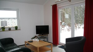 Un salon avec un téléviseur, une table basse en bois, un canapé noir, un fauteuil noir et une fenêtre avec des rideaux rouges.