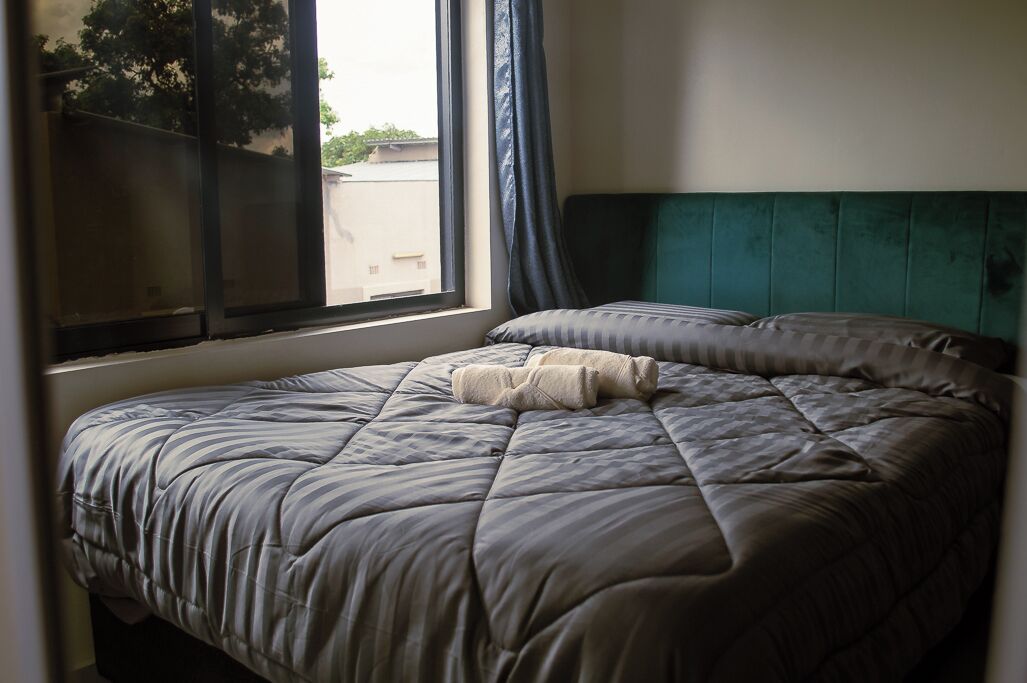 Uma cama com um cobertor listrado e uma toalha enrolada, uma janela com vista para edifícios, e uma cabeceira azul-esverdeada.