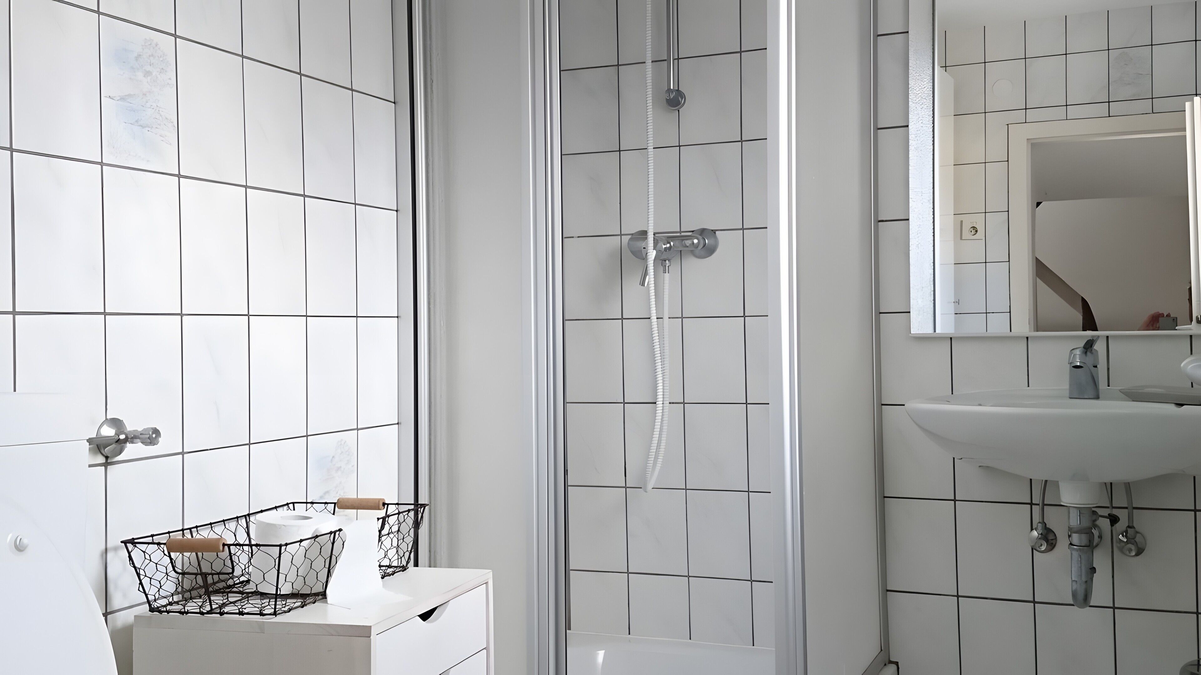 Ein Badezimmer mit weißen Fliesen, einer Duschkabine, einem Waschbecken mit Spiegel und einer Toilette mit Papierhandtuchhalter.