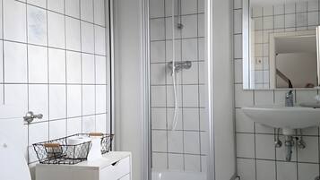 Ein Badezimmer mit weißen Fliesen, einer Duschkabine, einem Waschbecken mit Spiegel und einer Toilette mit Papierhandtuchhalter.