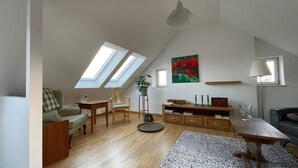 Ett rum i loftstil med ett sluttande tak, takfönster, ett matbord i trä, en soffa, en stol, ett sidobord, en golvlampa och en tavla.