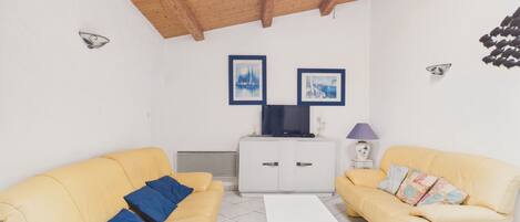 A living room with two yellow sofas, a white coffee table, a TV on a cabinet, and framed pictures on the wall.