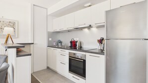 Una cuina moderna amb armaris blancs, un frigorífic d'acer inoxidable, un forn integrat i un taulell amb una paperera i utensilis.