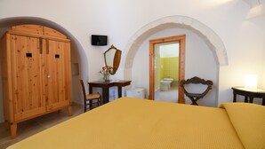A bedroom with a bed, wooden wardrobe, desk, chair, mirror, and an arched doorway leading to a bathroom.