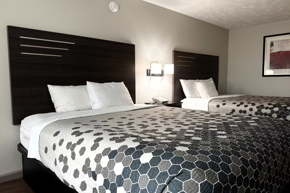 A hotel room with two beds, a dark headboard, a nightstand with a phone, and a wall-mounted light fixture.