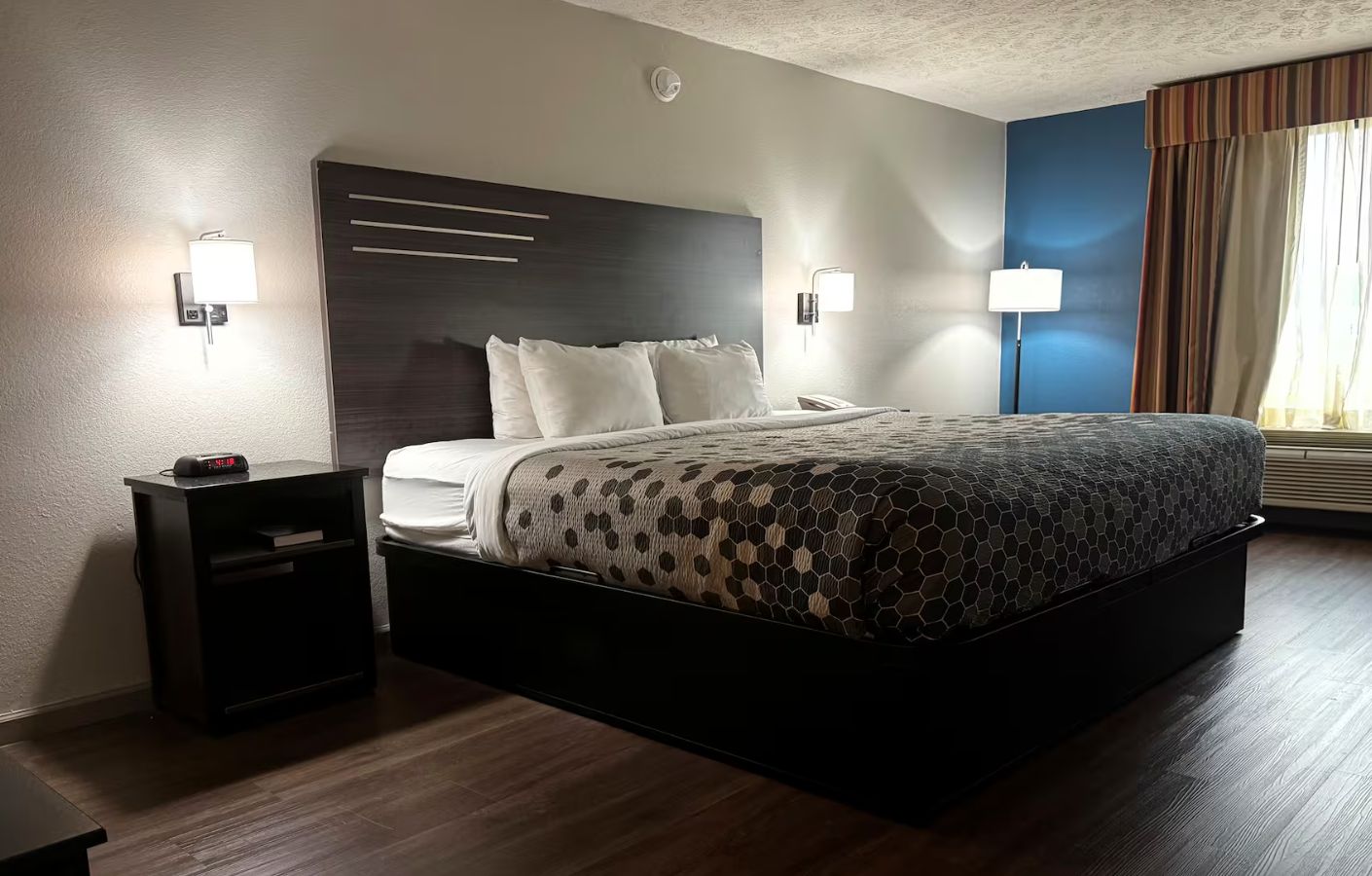 A hotel room with a large bed, bedside tables, a wall-mounted clock, and a window with curtains.