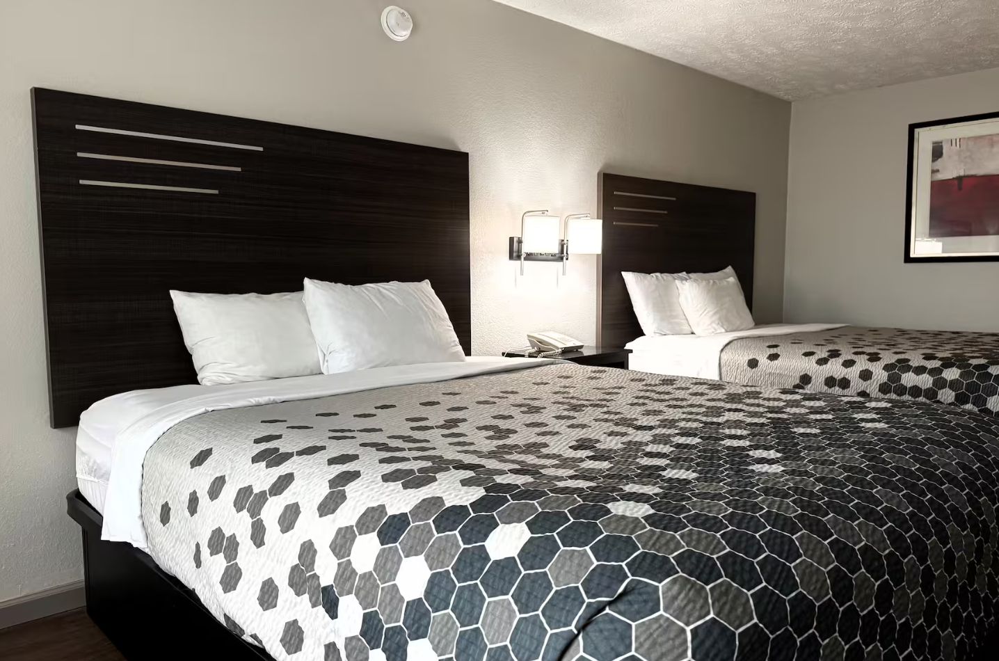 A hotel room with two beds, a dark headboard, a nightstand with a phone, and a wall-mounted light fixture.