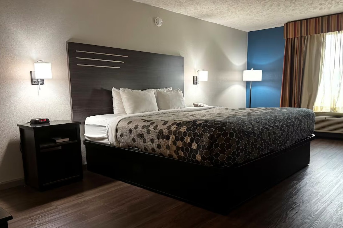 Habitación de hotel con una cama grande, mesitas de noche, un reloj en la pared y una ventana con cortinas.