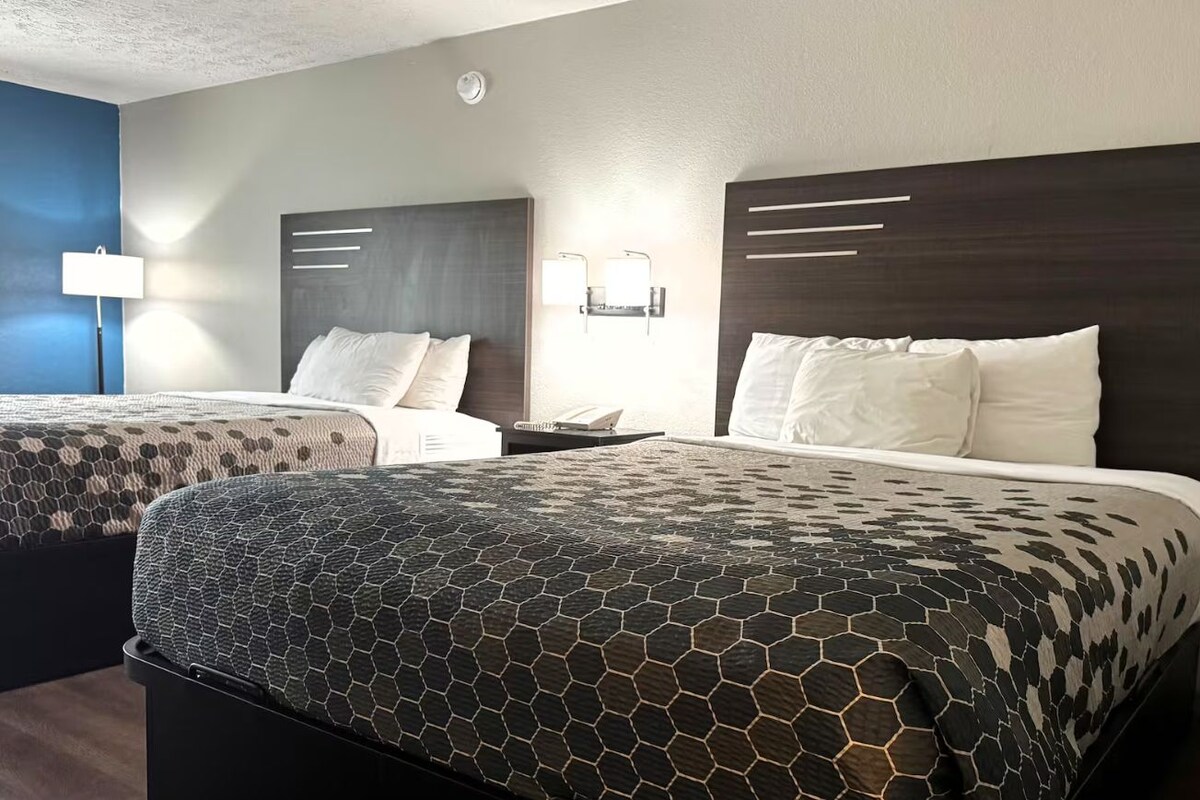 Habitación de hotel con dos camas, cada una con cabecero oscuro y ropa de cama blanca. Se observan lámparas de pared y una lámpara de techo.