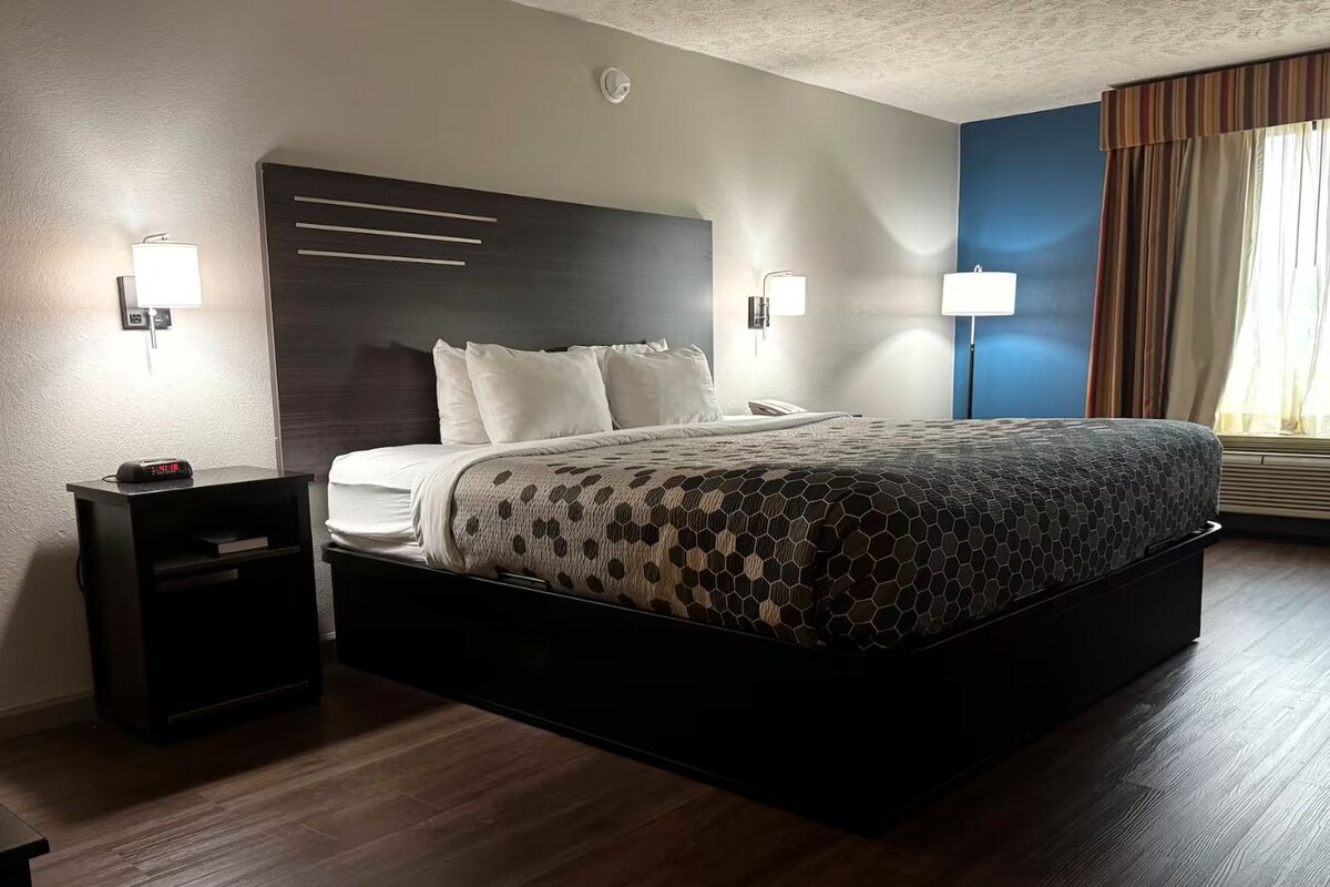 Habitación de hotel con una cama grande, mesitas de noche, un reloj en la pared y una ventana con cortinas.