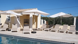 Un espace aménagé au bord de la piscine, avec des chaises longues et des parasols, une piscine carrelée et un bâtiment doté d’un toit en tuiles.