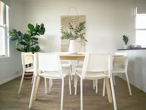 Un comedor con mesa de madera y sillas blancas, un colgador de macramé y una planta en maceta.