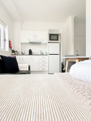Une chambre d’hôtel moderne et compacte, dotée d’une kitchenette, d’un lit avec une couvre-lit à rayures et donnant sur la chambre voisine.